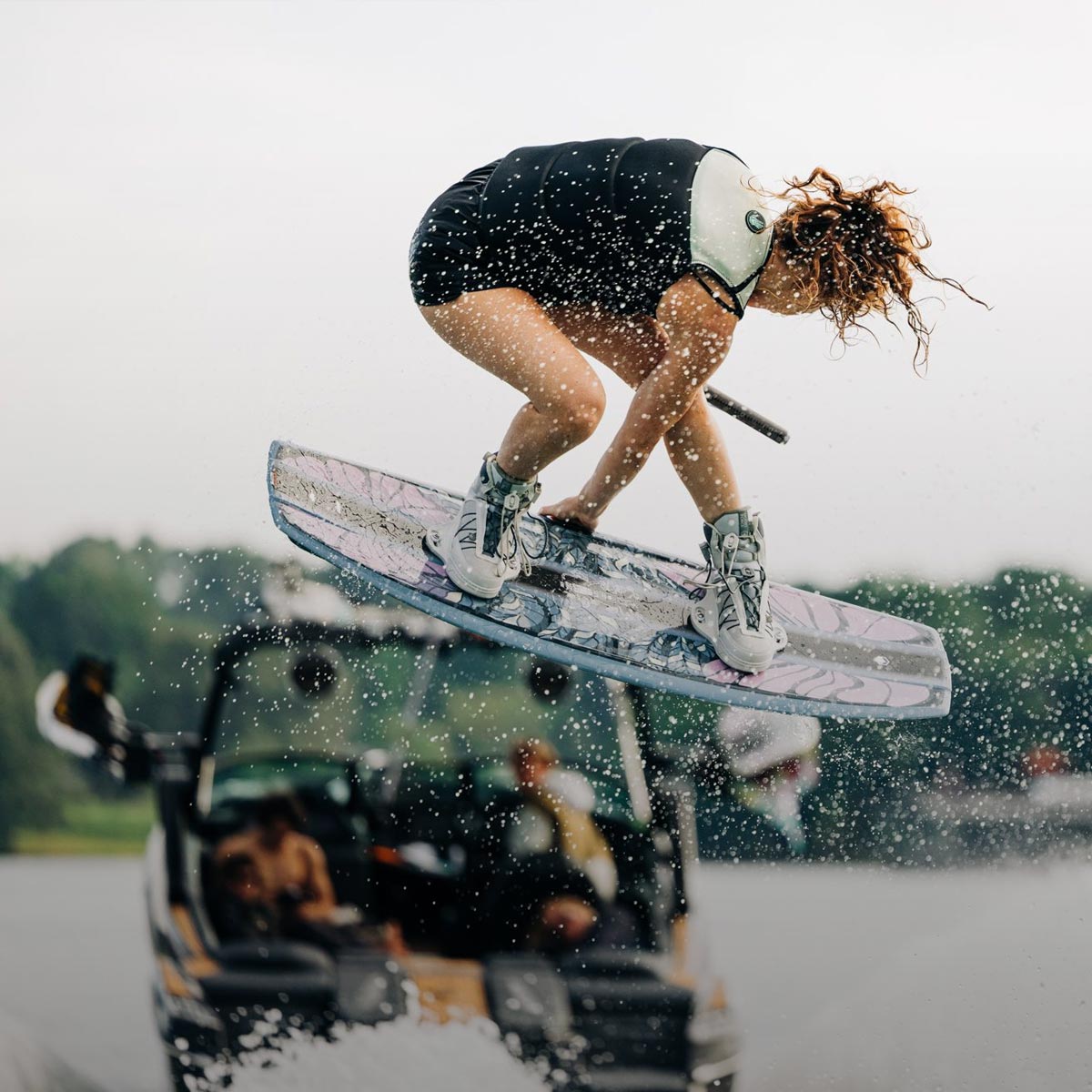 Liquid Force Boat Wakeboard Package Collection - Closeup shot of LF Athlete Meagan Ethell grabbing her board mid-air behind a wakeboard boat.